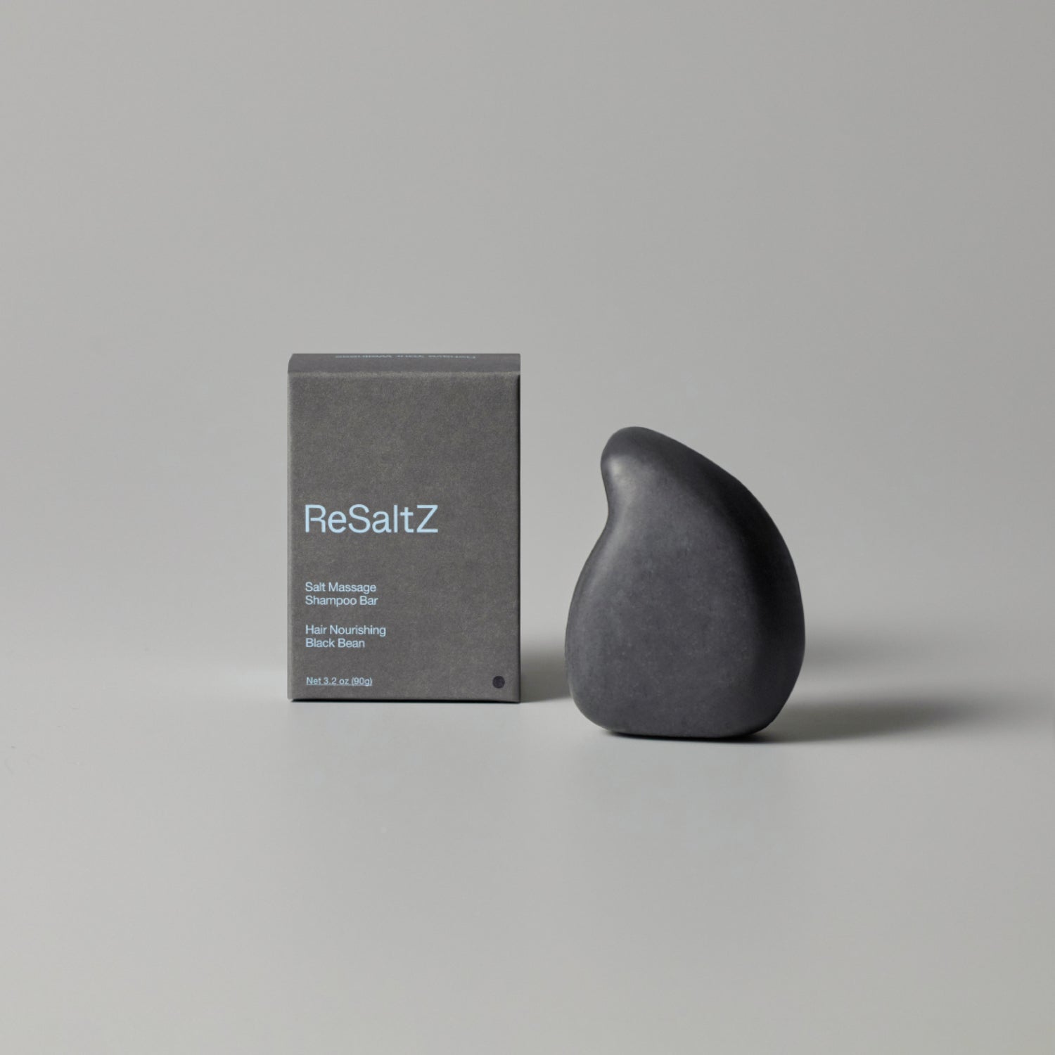 ReSaltZ Salt Massage Shampoo Bar in Black Bean, featuring a sleek, teardrop-shaped black shampoo bar next to its minimalist gray packaging. The product promotes hair nourishment and scalp care.