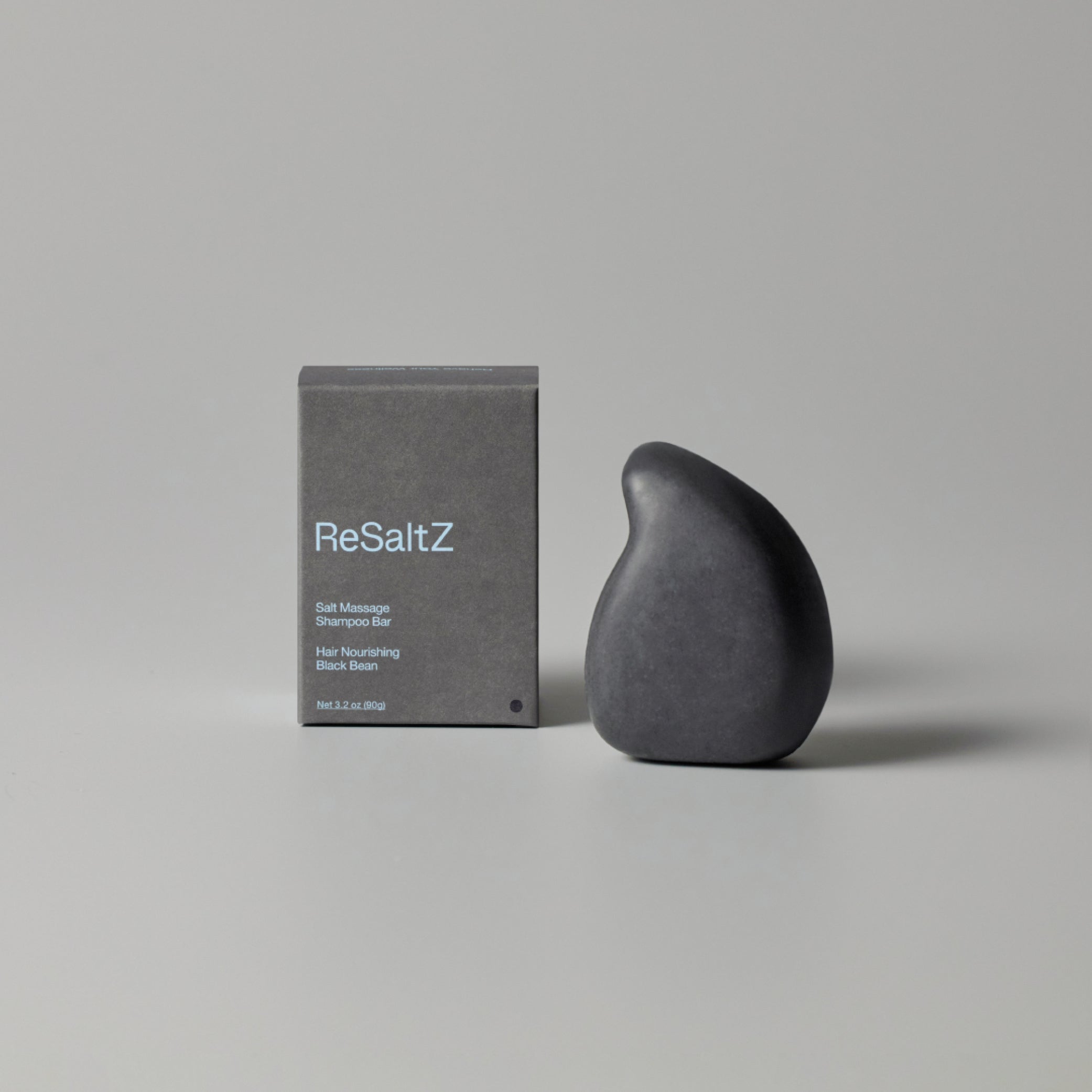 ReSaltZ Salt Massage Shampoo Bar in Black Bean, featuring a sleek, teardrop-shaped black shampoo bar next to its minimalist gray packaging. The product promotes hair nourishment and scalp care.